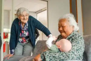 ‘Het Prins Claushuis voelt als familie’ 2 Een zorgmedewerkter is met haar pasgeboren baby op bezoek in Het Prins Claushuis. Een bewoonster heeft haar op de arm terwijl een andere bewoonster eroverheen gebogen staat en vertederd toekijkt.