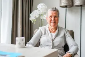 Bewoonster van Het Zevenheuvelenhuis in haar eigen studio, tussen haar vertrouwde spullen. Ze zit aan tafel en lacht in de camera