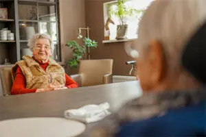 Twee bewoonsters van een zorglocatie van Dagelijks Leven tegenover elkaar aan tafel. Een van hen wordt over de schouder gefotografeerd, de ander lacht in de camera. Ook in Hardenberg kunnen mensen met dementie bij Dagelijks Leven terecht. 