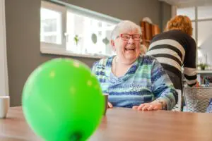 Een bewoner aan tafel in een woonzorglocatie van Dagelijks Leven. Ze moet lachen en kijkt opzij. voor haar een groene ballon. Mensen met dementie kunnen ook in Heerhugowaard bij Dagelijks Leven wonen. 