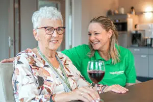 Een bewoonster van Dagelijks Leven zit aan tafel in de huiskamer van zorgorganisatie Dagelijks Leven in Deurne, voor mensen met dementie. Ze drinkt een glaasje wijn. Naast haar zit een medewerker, die naar haar lacht. 