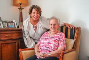 Een bewoonster samen met haar dochter samen in haar studio in een woonzorglocatie voor mensen met dementie van Dagelijks Leven. Ze zit voor een kast waarboven op allemaal fotolijstjes staan. 