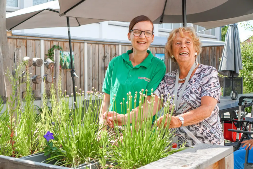 Een bewoonster met Alzheimer en een verzorgende IG staan buiten in de tuin van het verzorgingshuis voor mensen met dementie. Ze werken in de kruidentuin en lachen naar de camera.