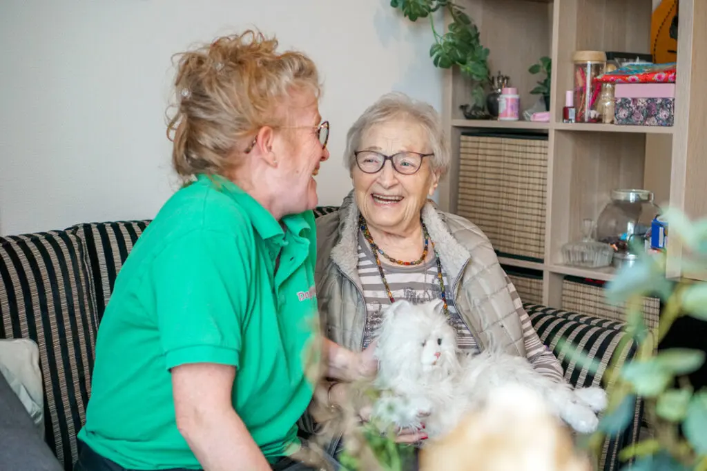 Een bewoonster met Alzheimer kletst met een zorgmedewerkster van Dagelijks Leven in haar studio in het verzorgingshuis voor mensen met dementie. Op haar schoot zit een Robot Kat