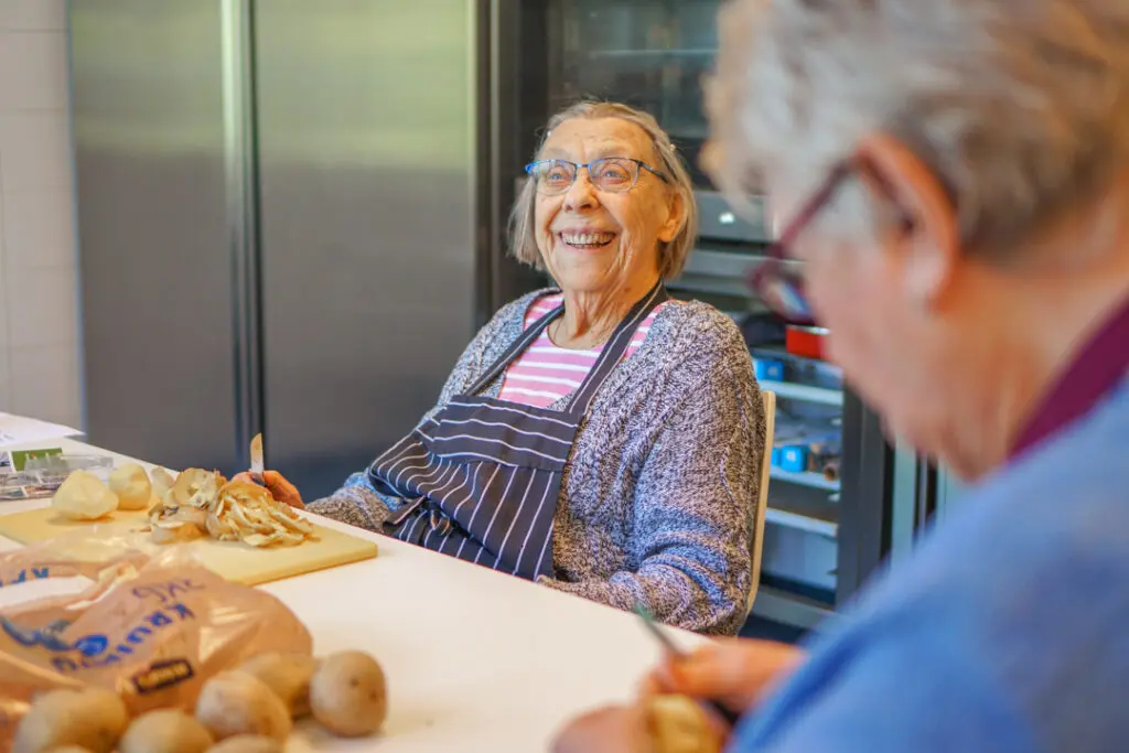 Een bewoonster van de kleinschalige zorglocatie voor mensen met dementie lacht naar de camera tijdens het schillen van de aardappels