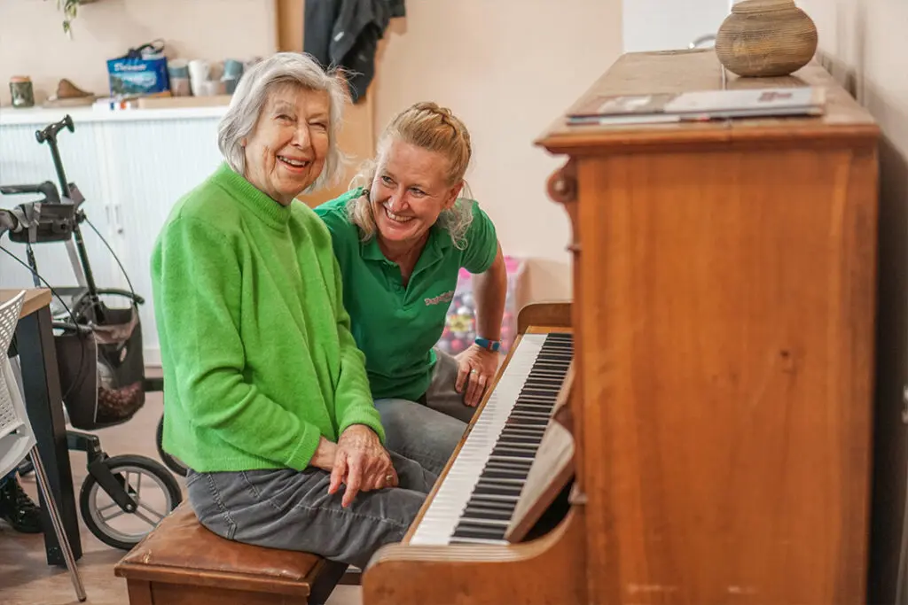 Een bewoner en een medewerker van Dagelijks Leven kijken lachend in de camera. Ze zitten samen bij een piano. Ter illustratie bij informatie over kleinschalig en betaalbaar wonen met dementie bij Dagelijks Leven.