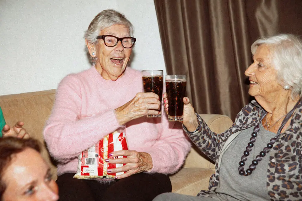 Twee bewoners met dementie proosten met elkaar terwijl ze een feestje hebben met de verzorgende ig, verpleegkundige en helpende van de zorglocatie van Dagelijks Leven tijdens de opnames voor de Lekker met de meiden-campagne