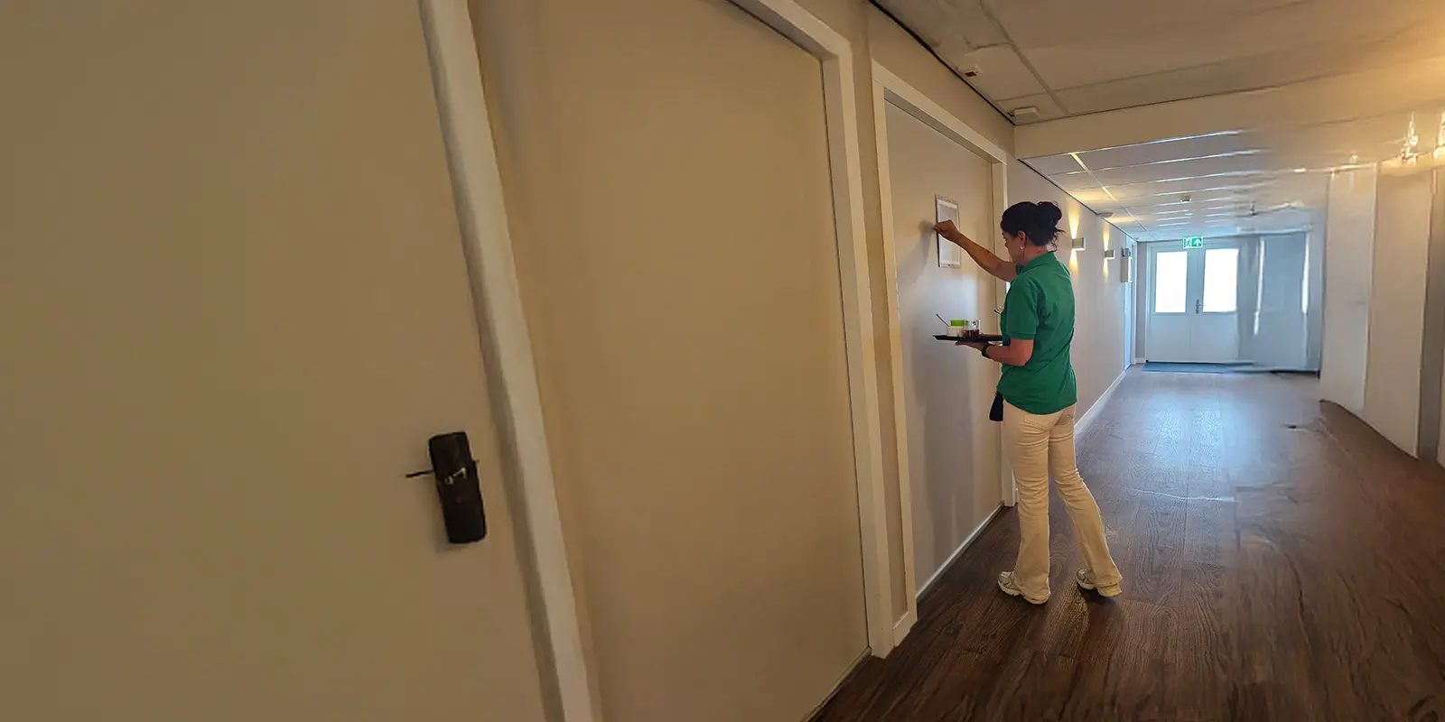 Gespecialiseerd verzorgende psychogeriatrie klopt op een studiodeur van de bewoner van een kleinschalige woonzorglocatie voor mensen met dementie. Ze brengt ontbijt op bed. Foto bij een blog over hoe je als zorgmedewerker goed voor ogen moet houden dat gedrag veroorzaakt kan worden door de ziekte, niet door de persoon zelf. In dit geval vasculaire dementie.