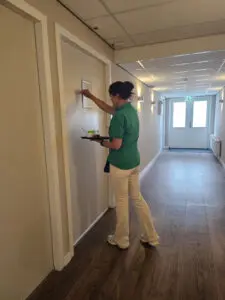 Gespecialiseerd verzorgende psychogeriatrie klopt op een studiodeur van de bewoner van een kleinschalige woonzorglocatie voor mensen met dementie. Ze brengt ontbijt op bed. Foto bij een blog over hoe je als zorgmedewerker goed voor ogen moet houden dat gedrag veroorzaakt kan worden door de ziekte, niet door de persoon zelf. In dit geval vasculaire dementie.