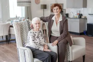 Foto van dochter met moeder, die in Het RIngbaanhuis woont, onze kleinschalige woonzorglocatie voor mensen met dementie in Tilburg. Moeder zit in fauteuil, dochter op de stoelleuning.