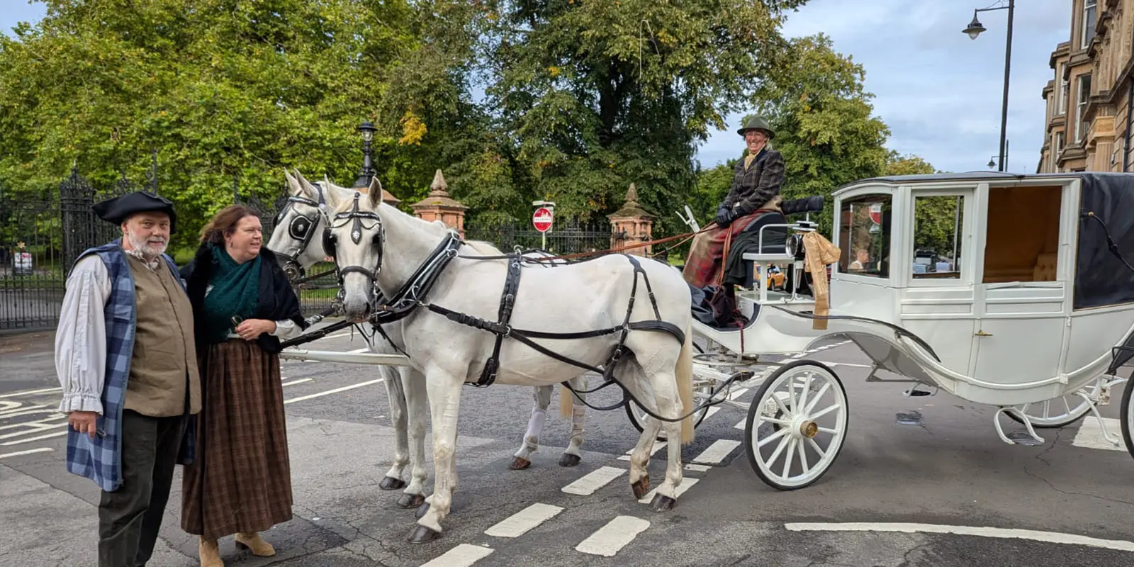 Op de foto is een witte koets te zien, met op de koets een vrouw. Voor de koets staan twee witte paarden met daarnaast een man en een vrouw in 18e eeuwse kleding.