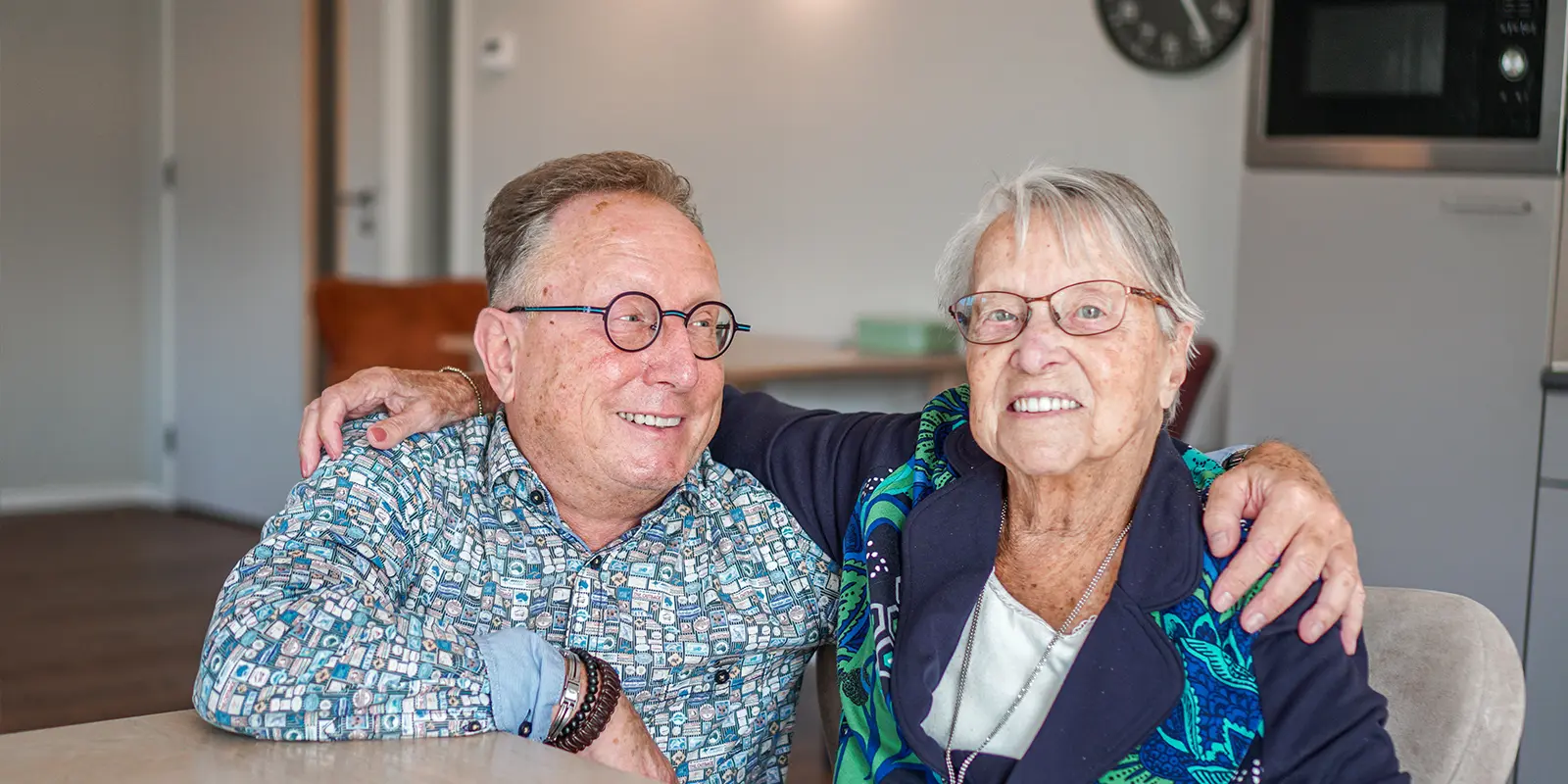 Foto van zoon en moeder, aan tafel in de huiskamer van Het Stappegoorhuis, onze kleinschalige woonzorglocatie voor mensen met dementie in Tilburg. Moeder heeft arm om zoon heen geslagen en kijkt lachend in de camera. Zoon kijkt lachend naar zijn moeder.