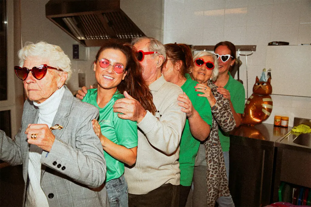 Drie zorgmedewerkers van Dagelijks Leven doen de polonaise voor de campagne "Lekker met de meiden" samen met drie bewoners met dementie. De medewerkers zijn verpleegkundige, verzorgende ig of helpende