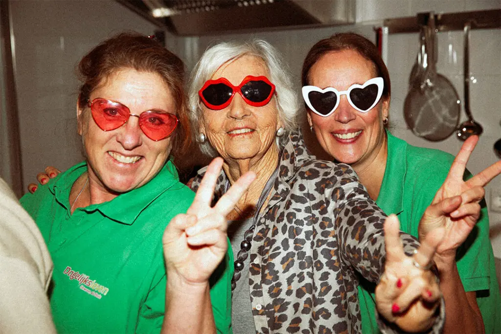 Twee medewerksters en een bewoonster met dementie lachen naar de camera en hebben bijzondere brillen op. Ze poseren voor de wervingscampagne voor "Lekker met de meiden", waarin Dagelijks Leven zoekt naar een nieuwe verzorgende IG, verpleegkundige of helpende