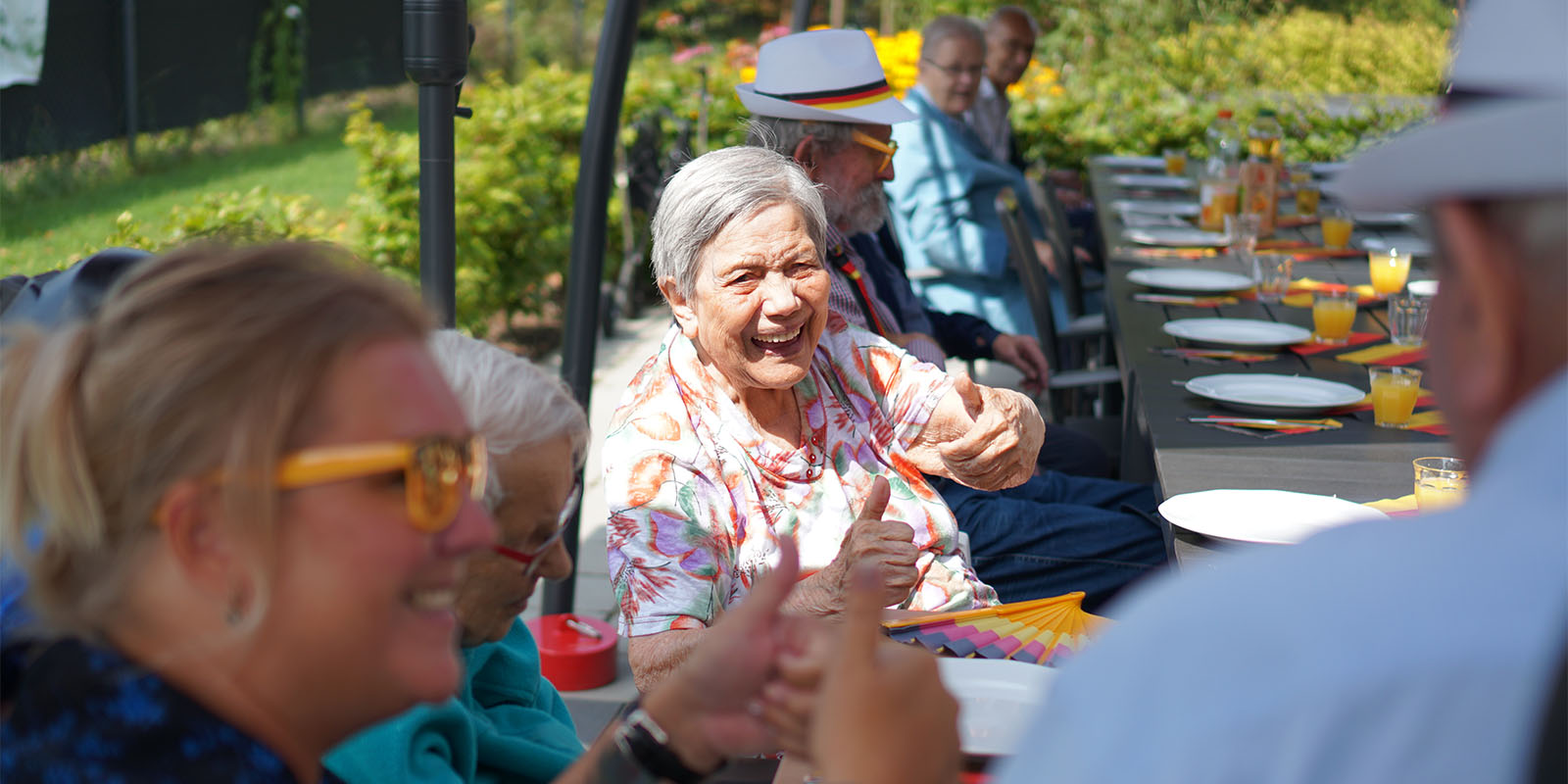 Bewoners Dagelijks Leven zitten samen aan tafel op het terras in de zon. De bewoner in het midden moet heel har lachen.