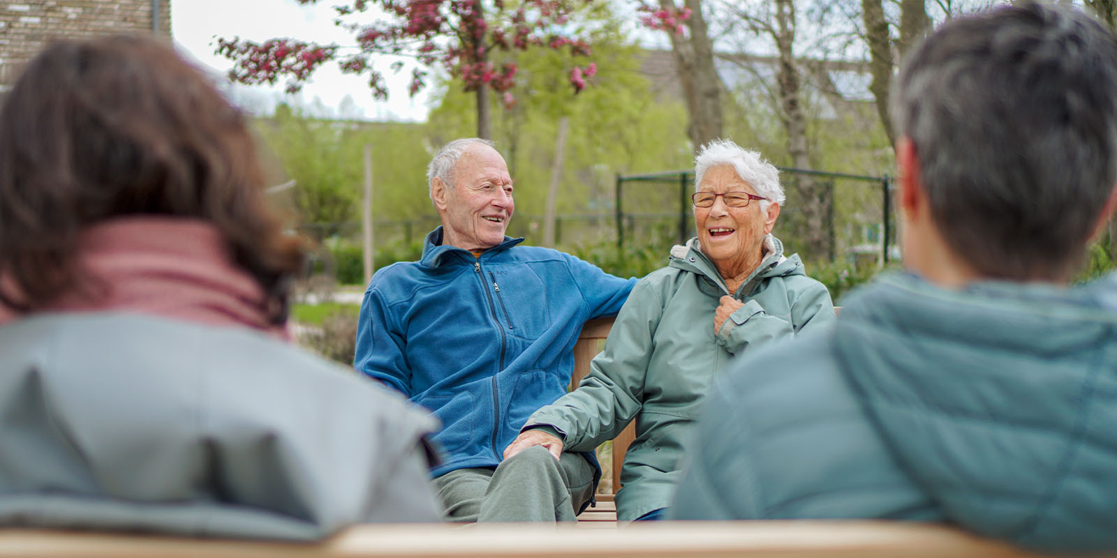 Echtpaar met dementie dat samenwoont in de echtparenstudio van Dagelijks Leven woont samen buiten op een bankje, lachend. Hun twee dochters kijken toe.