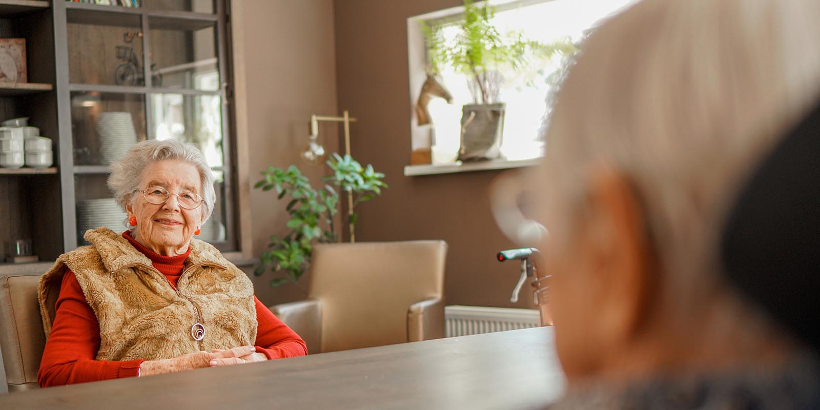 Bewoonster met dementie zit tegenover een andere bewoner aan tafel in een woonkamer van Dagelijks Leven