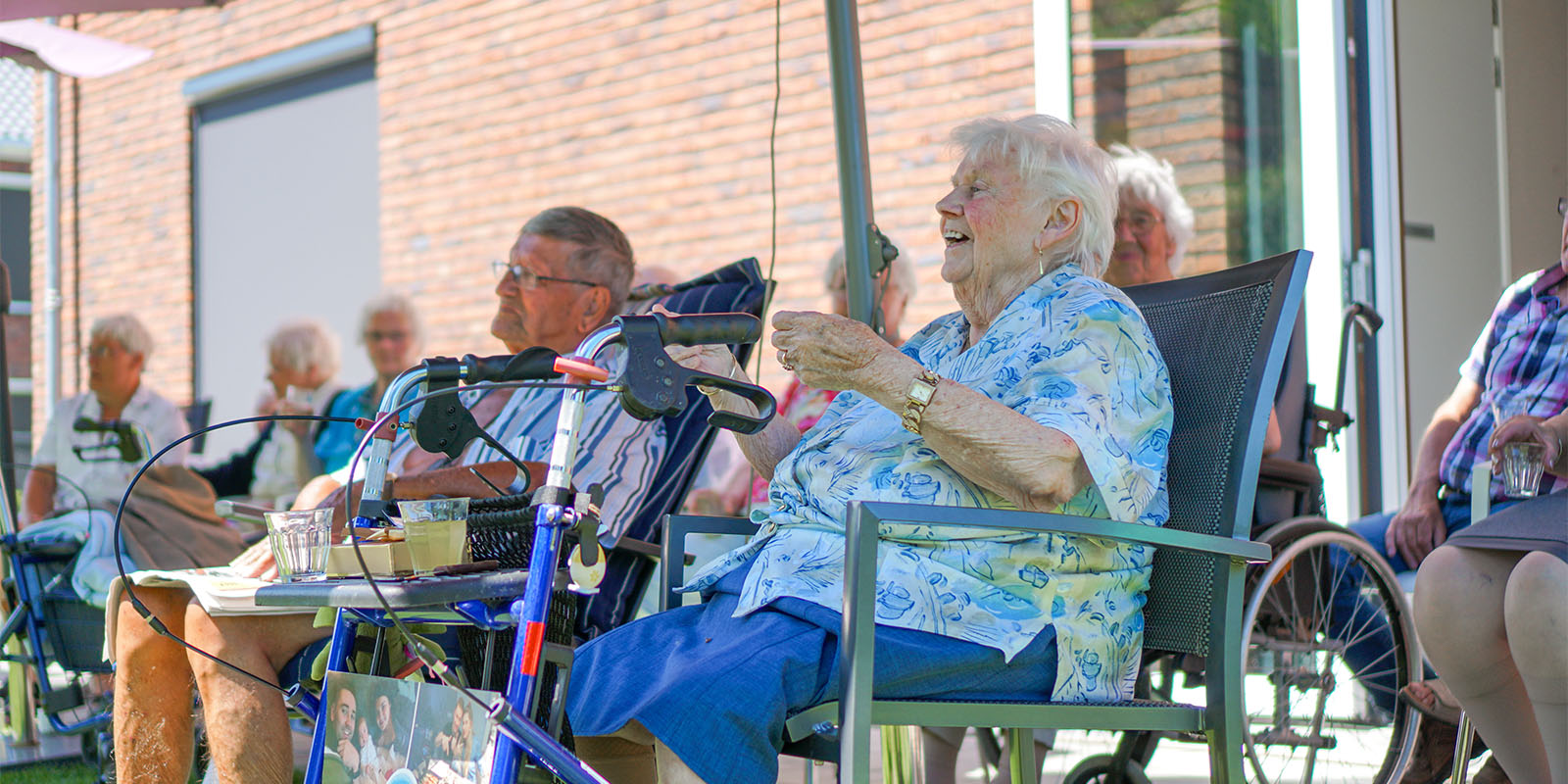 Foto ter illustratie bij het blog van specialist ouderengeneeskunde. Op de foto zit een aantal bewoners buiten in de zon, ze lijken te genieten van een voorstelling.