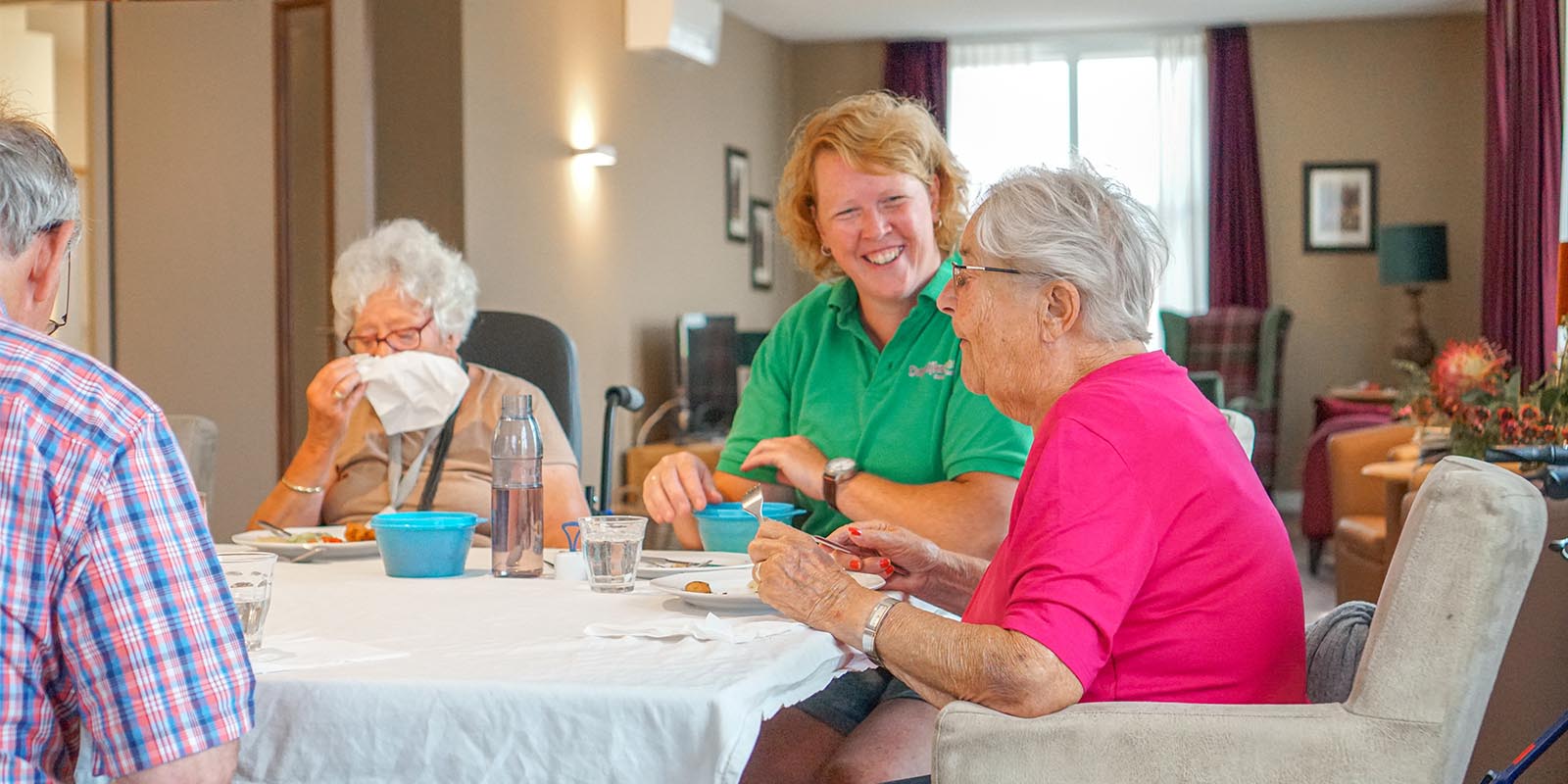 Bewoners met dementie genieten samen van de maaltijd aan een gedekte tafel. Ook de zorgmedewerker is aangeschoven. Rust, sfeer en beleving: een fijne maaltijd voor mensen met dementie.