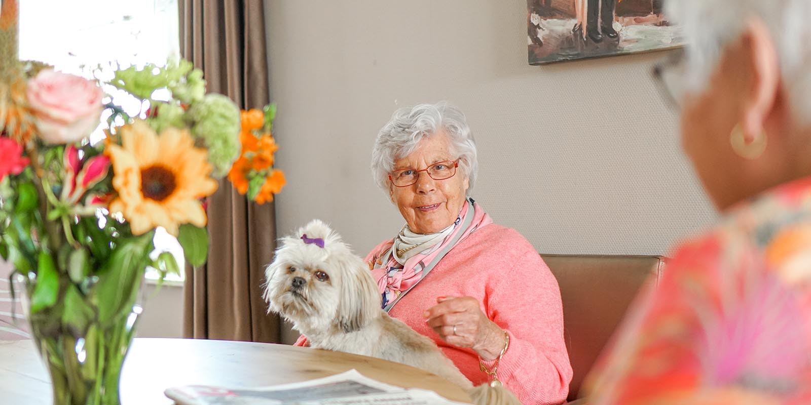 Wonen met zorg voor mensen met dementie in Venray: dat is Dagelijks Leven. Op de foto een bewoonster aan tafel, lachend, met een hondje op schoot.