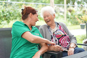 Een verpleegkundige zit met een bewoner met dementie in de tuin van een kleinschalige woonzorglocatie. Ze lachen naar elkaar.