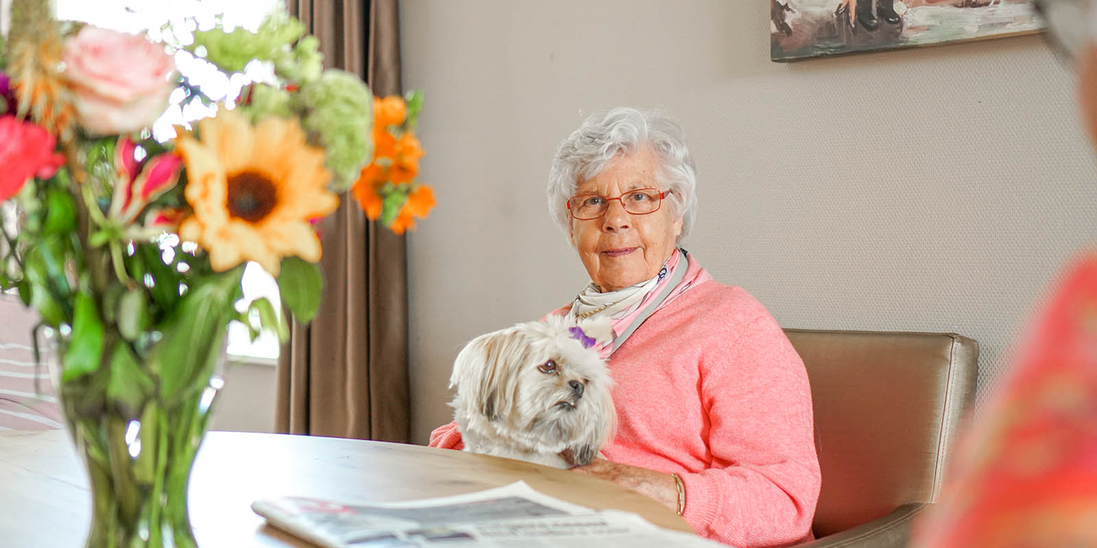 Een bewoonster van een kleisnchalige woonvorm voor mensen met dementie zit aan tafel en knuffelt met een hondje.
