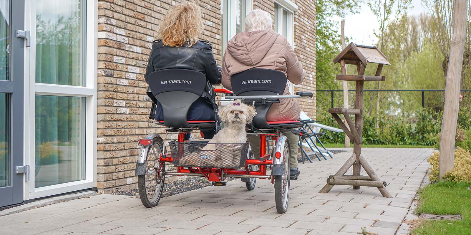 Een bewoner met dementie samen met haar dochter op een duofiets, van achter gefotografeerd terwijl ze wegrijden. Achter in het bakje zit een klein hondje dat de camera inkijkt.