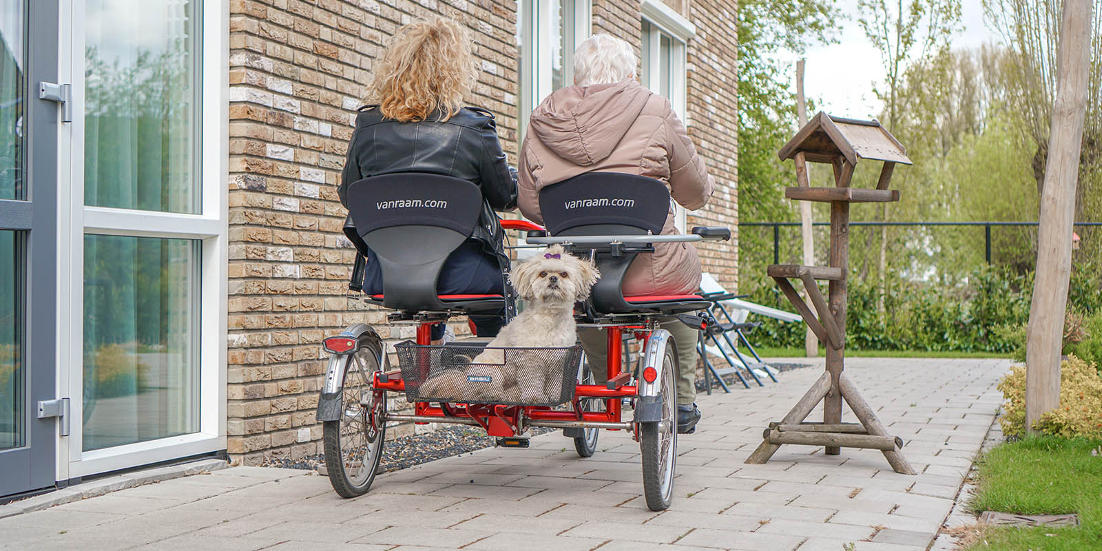 Aan Het Enkhuis, kleinschalig wonen voor mensen met dementie, is een duofiets geschonken. Foto ter illustratie: we zien van achteren een moder met dementie samen met haar dochter wegfietsen op de duofiets. In het bakje achterop de fiets het hondje van de dochter, die de camera in kijkt.
