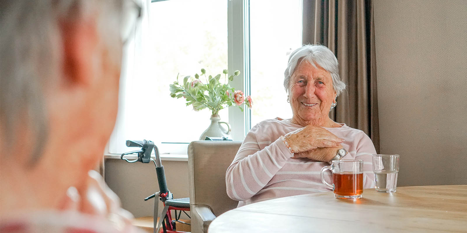 Foto van een bewoonster aan tafel, met een glas thee, in een huis van Dagelijks Leven. Ter illustratie bij een blog van psycholoog Ribanna, die meer vertelt over haar werk met mensen met dementie.