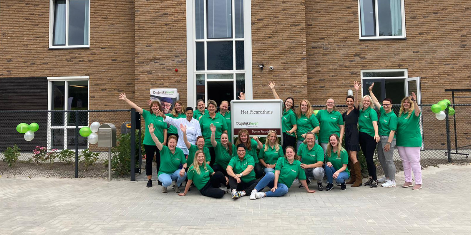 Foto van het team tijdens de opening van Het Picardthuis, kleinschalige woonvorm voor mensen met dementie in Coevorden. In hun kenmerkende groene polo's staan ze voor het huis, met feestelijk hun armen in de lucht.