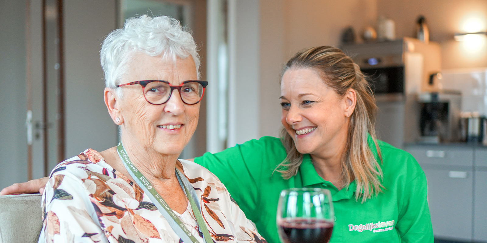 Een bewoonster van een huis van Dagelijks Leven zit aan de eettafel samen met een verzorgende. De bewoonster proost met een glas wijn en ze lachen naar elkaar. Dagelijks Leven bouwt ook twee woonzorglocaties voor mensen met dementie in Sittard.