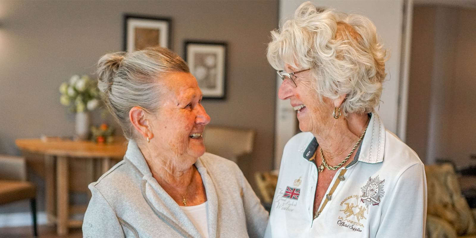 Zussen lachen breeduit naar elkaar tijdens een onderonsje in de huiskamer van Het Zevenheuvelenhuis, zorglocatie voor mensen met dementie in Groesbeek.