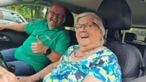 GVP Pieter samen met een bewoonster met dementie in de auto. Ze lachen en Pieter steekt een duim op. Ze gaan samen op pad om de hartenwens van de bewoonster te vervullen: een keer terug naar haar oude buurt.