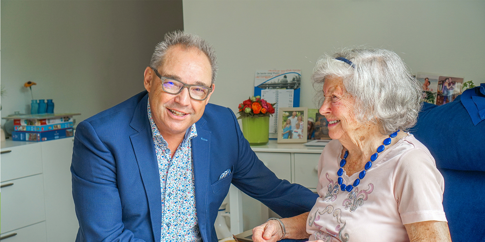 Specialist ouderengeneeskunde op bezoek bij een bewoner met dementie van Dagelijks Leven, kleinschalige woonzorglocatie. Hij lacht naar de camera, de bewoonster lacht naar de specialist, die onderdeel is van de vakgroep Advies, Behandeling en Begeleiding.