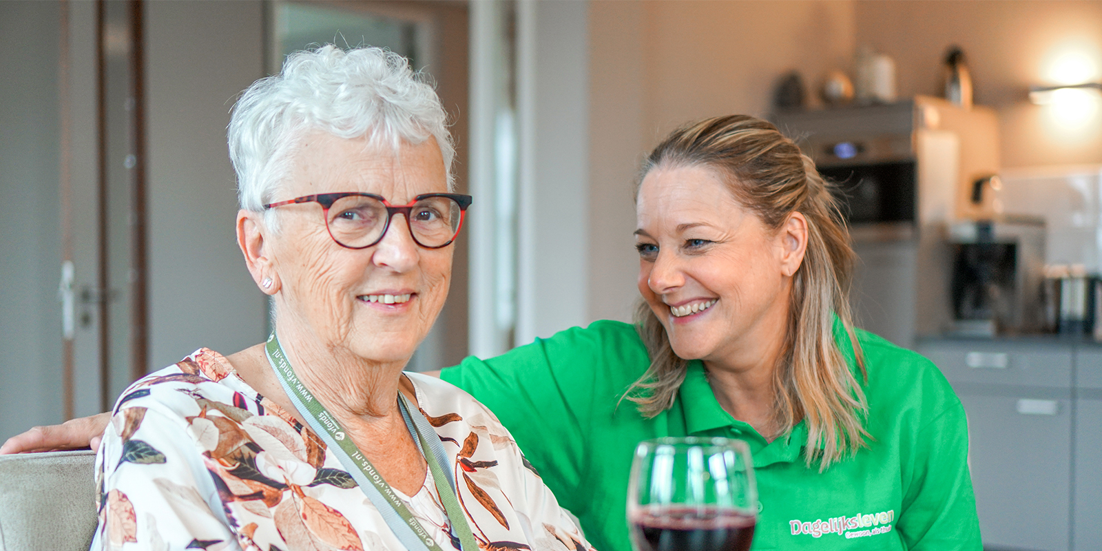 Een bewoonster met dementie samen met een medewerkster aan tafel in een kleinschalige zorglocatie van Dagelijks Leven. De medewerker lacht naar de bewoner, die met een glaasje wijn lachend de camera inkijkt. In Hardenberg zijn twee locaties van Dagelijks Leven.