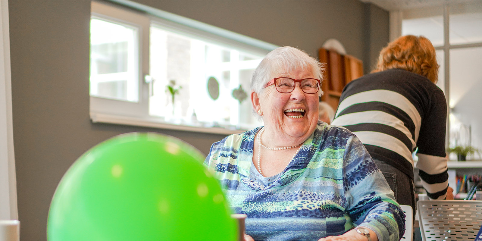 Humor houdt je jong, ook als je eigenlijk al wat ouder bent, weet de activiteitenbegeleider van een huis van Dagelijks Leven voor mensen met dementie. Op de foot zien we een bewoonster heel hard lachen. Ze zit aan tafel, voor haar een groene ballon.