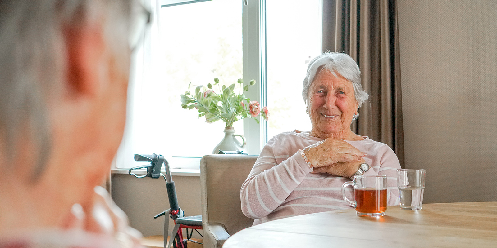 Bewoner aan tafel in de huiskamer van Dagelijks Leven, met een kopje thee. ze lacht naar de camera. Dagelijks Leven heeft in Deurne drie kleinschalige zorglocaties voor mensen met dementie.