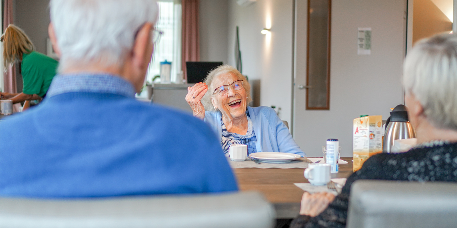 Een bewoonster zit lachend aan tafel in een woonzorglocatie van Dagelijks Leven, voor mensen met dementie. Ter illustratie bij een blog van verpleegkundig specialist Willy Erdhuizen over het belang van maaltijdbeleving in de ouderenzorg.