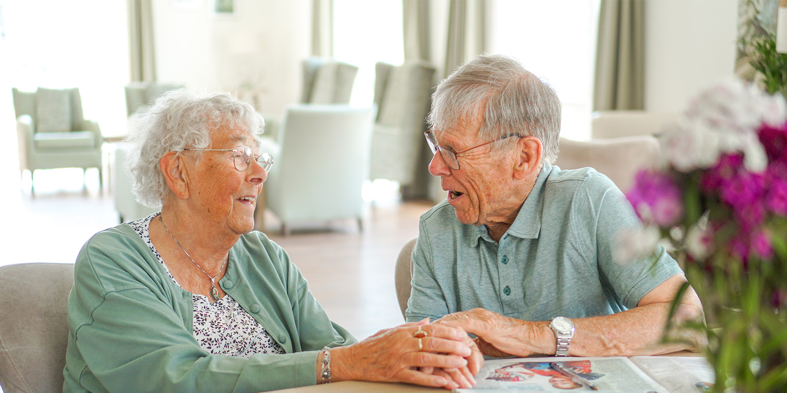 Herman en Joke wonen als echtpaar samen in Het Snijdelhuis in Boskoop, woonzorglocatie voor mensen met dementie. Op de foto zitten ze aan tafel in de huiskamer en lachen ze naar elkaar.