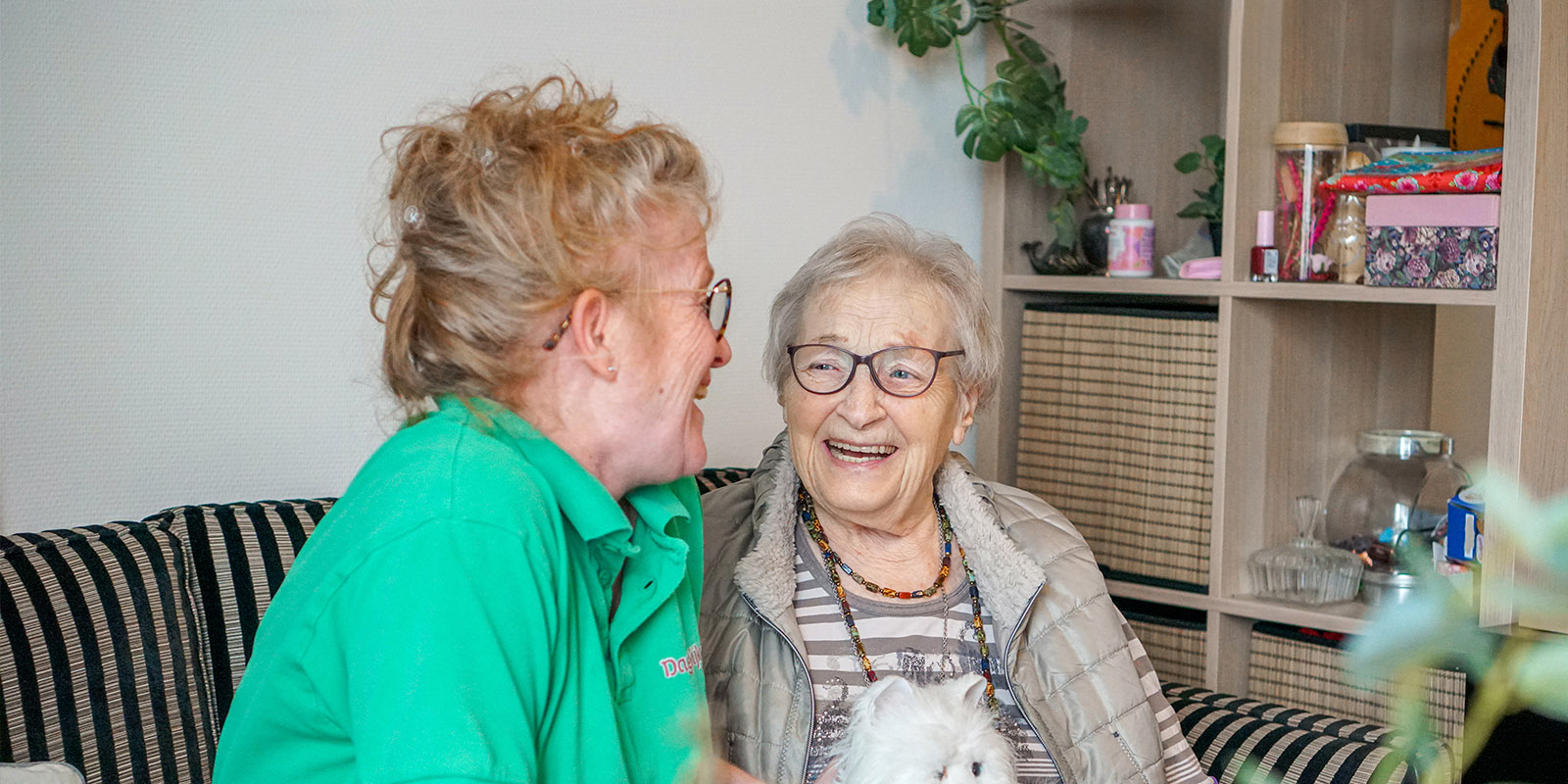 Een medewerkster van Dagelijks Leven zit samen met een bewoonster lachend op de bank in haar studio in de woonzorglocatie voor mensen met dementie. Bij Dagelijks Leven is zorg altijd aanwezig.