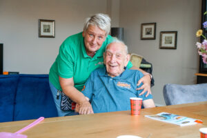 Een medewerkster van Dagelijks Leven lachend op de foto samen met een bewoner in een rolstoel. In onze woonzorglocaties voor mensen met dementie is zorg altijd aanwezig. 