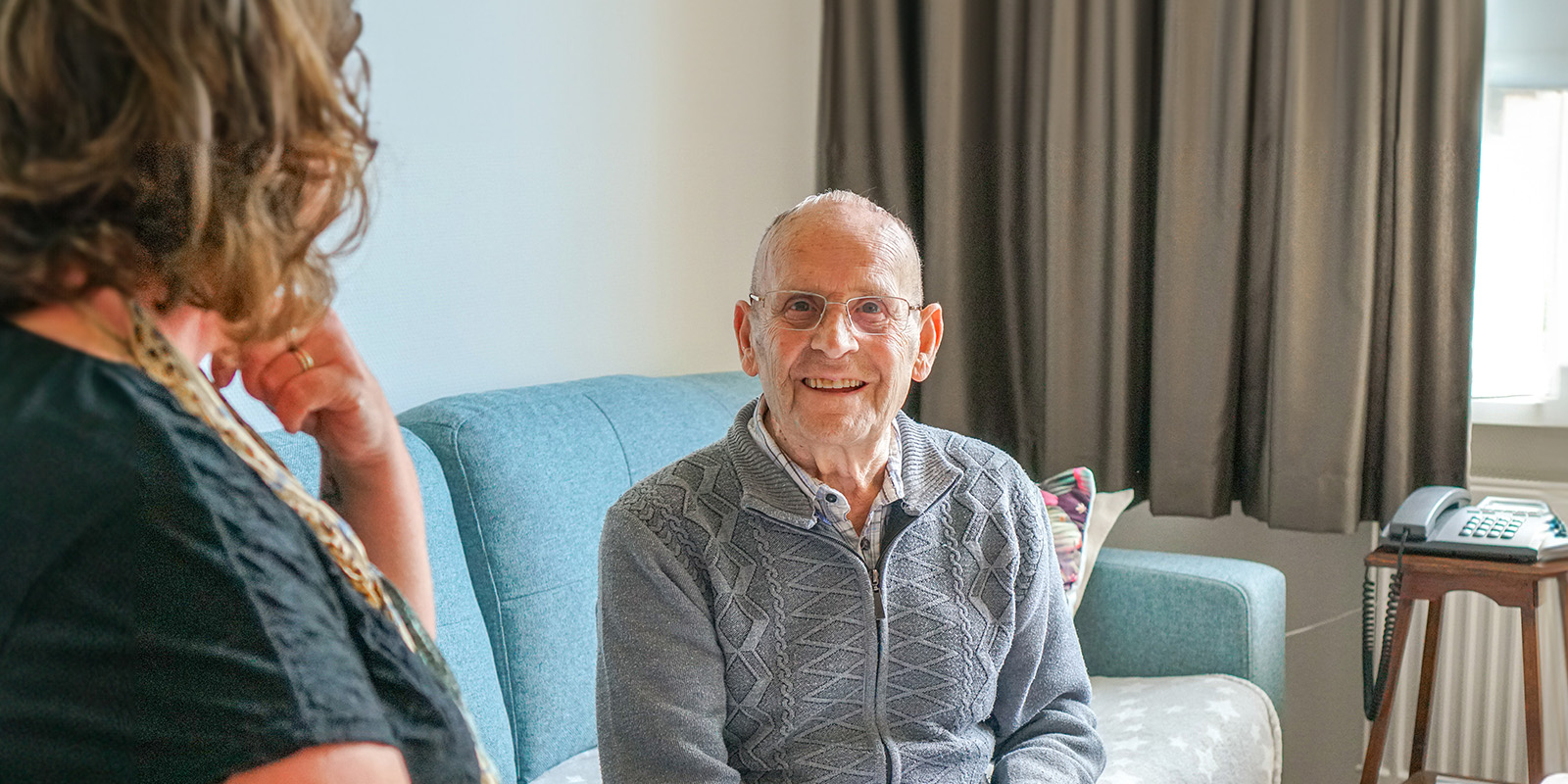 Een bewoner zit samen met zijn dochter in zijn studio in een woonzorglocatie voor mensen met dementie van Dagelijks Leven. Hij zit op de bank, zij op een stoel en ze kijken samen een oud fotoboek door.