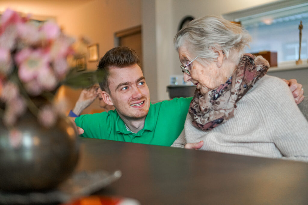 Een medewerker en een bewoonster met alzheimer kletsen met elkaar aan de eettafel van een verzorgingshuis van Dagelijks Leven