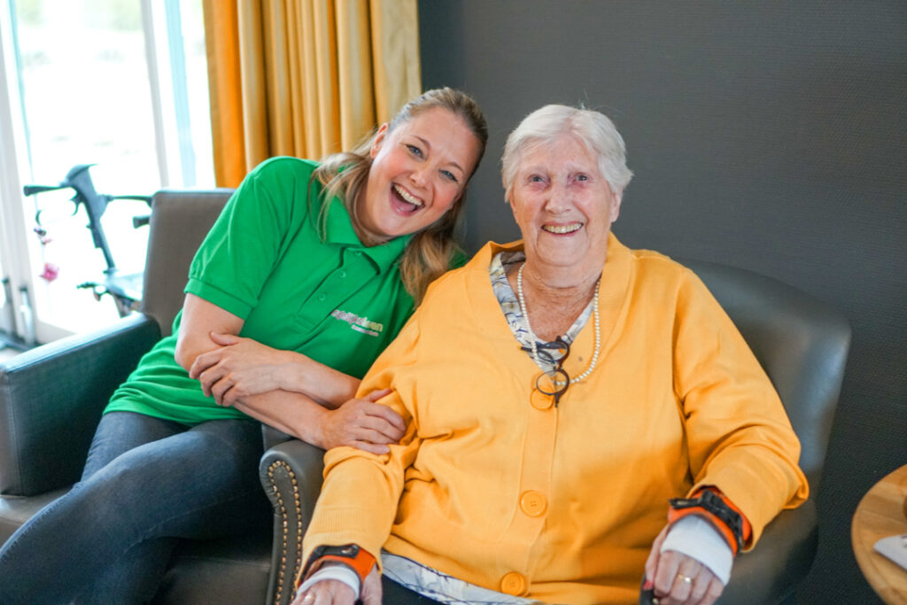 Een bewoonster met Alzheimer en een zorgmedewerkster zitten samen in een stoel van de zorglocatie voor mensen met dementie en lachen naar de camera