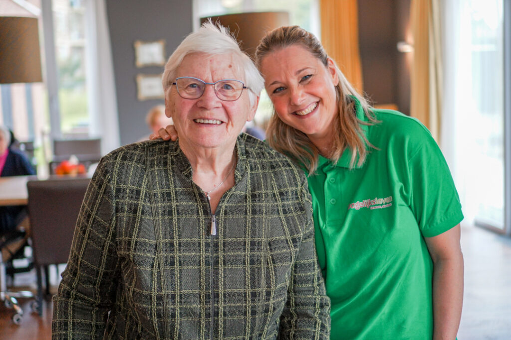 Een bewoonster met Alzheimer en een zorgmedewerkster van Dagelijks Leven lachen naar de camera in de woonkamer van de zorginstelling voor mensen met dementie