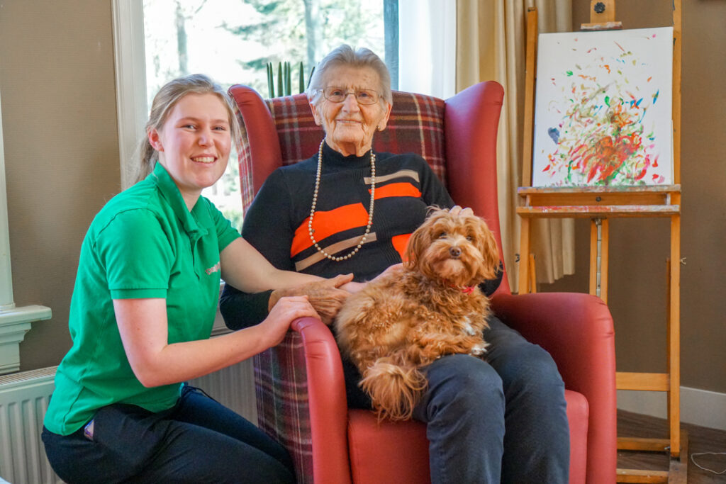 Een bewoonster met de ziekte van Alzheimer en een zorgmedewerkster van Dagelijks Leven knuffelen samen met een hondje dat op bezoek is in het verzorgingshuis