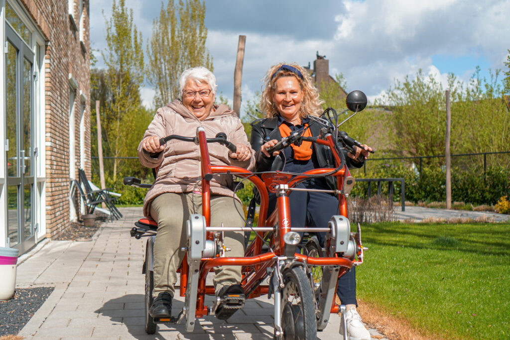 Een bewoonster met de ziekte van Alzheimer en een vrijwilliger fietsen samen op de duofiets in het verzorgingshuis van Dagelijks Leven.