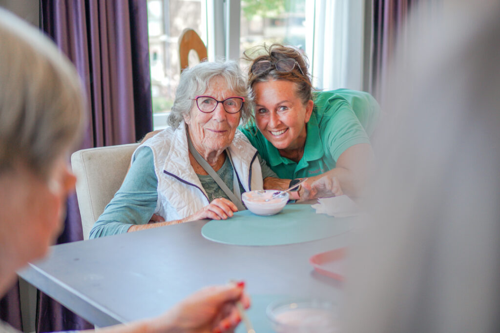 Een bewoonster met Alzheimer en een medewerkster kijken vrolijk de camera in tijdens het eten van een toetje in het verzorgingshuis voor mensen met dementie van Dagelijks Leven