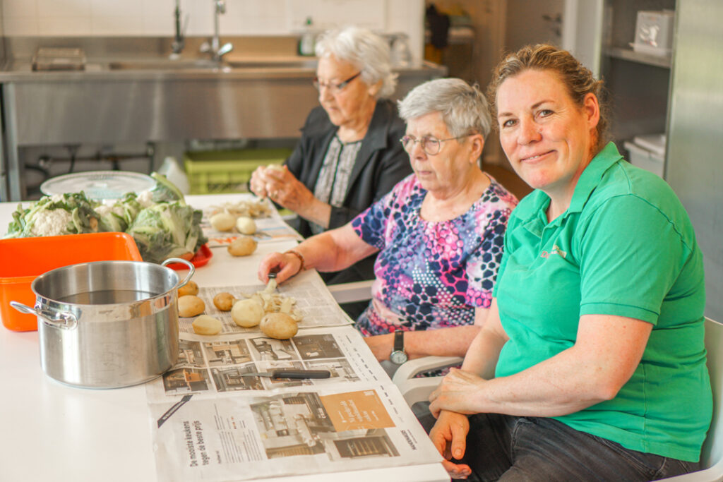 Een bewoonster met alzheimer en een bewoonster met vasculaire dementie helpen de kok mee om aardappels te schillen in de kleinschalige zorglocatie voor mensen met dementie.