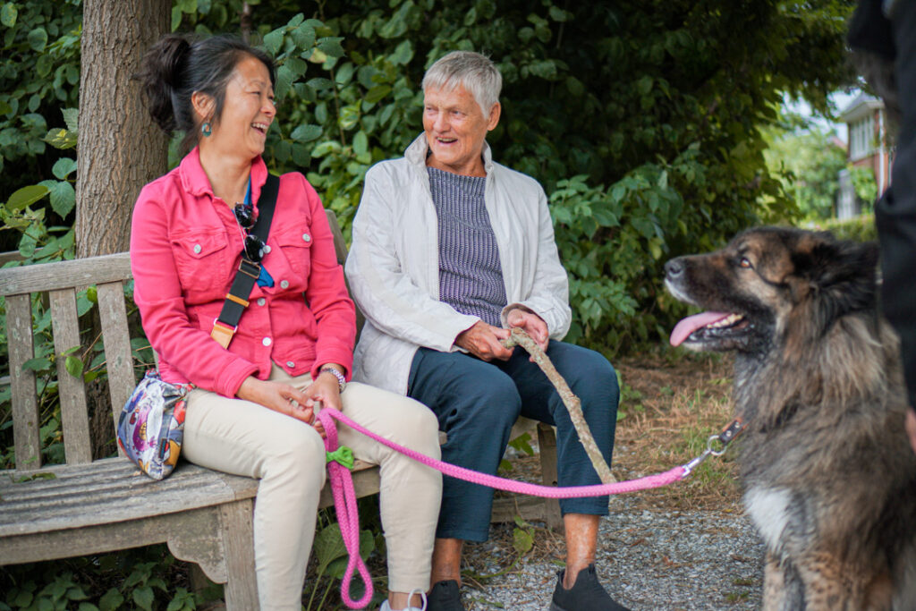 Een vrijwilligster van de kleinschalige zorglocatie van Dagelijks Leven wandelt samen met een bewoonster met Alzheimer een rondje met de hond. Samen zitten ze even op het bankje om uit te rusten.